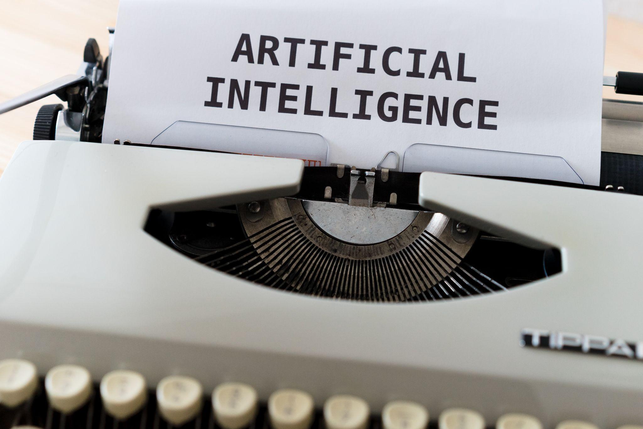 A white typewriter with paper in it bearing the words "ARTIFICIAL INTELLIGENCE" in black font.