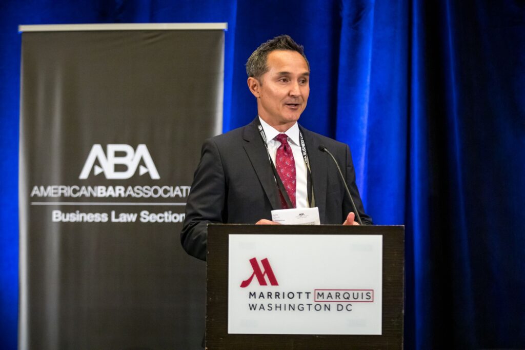 A man with salt-and-pepper hair in a black suit, white shirt, and patterned red tie speaks from a lectern, in front of a vibrant blue curtain. To the left behind him is a black banner with white text that reads, "American Bar Association Business Law Section" and displays the ABA logo.