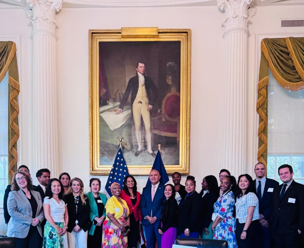 A diverse group of almost 20 people pose in front of a painting.