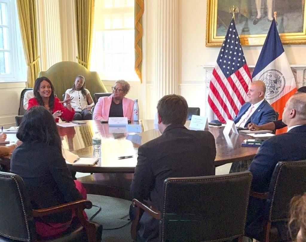 A woman in a red blazer speaks to multiple people in business attire seated around a round table, with more people seated around the edges of the formally decorated room.