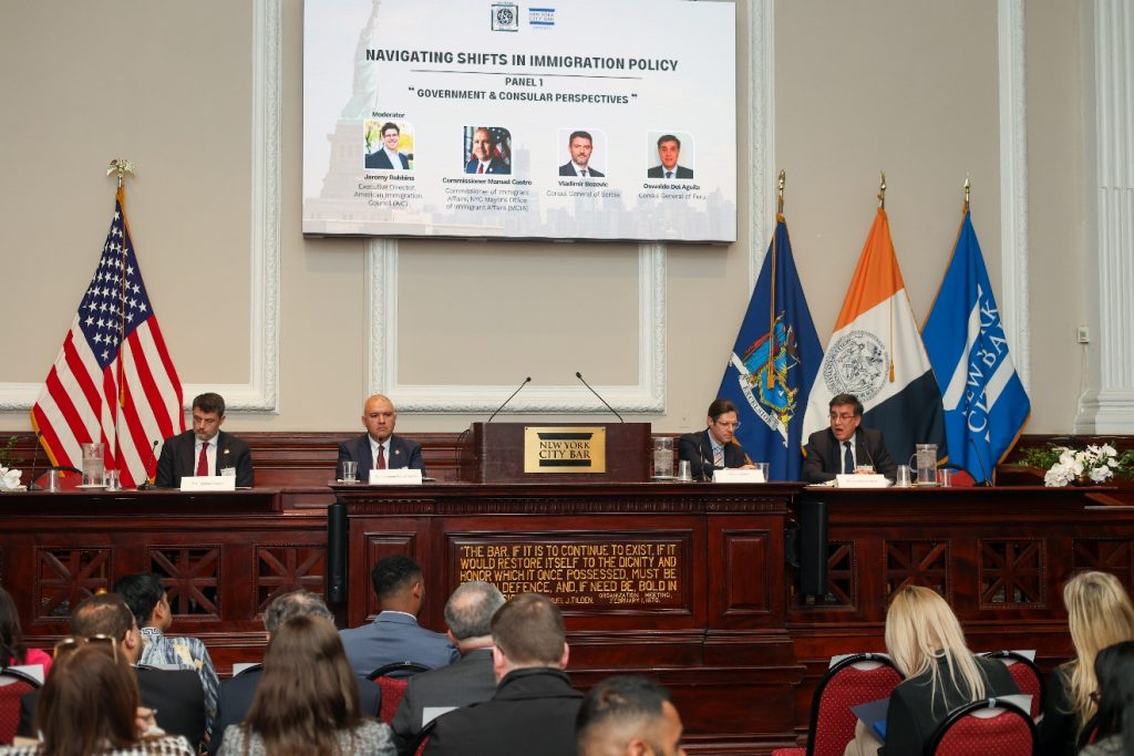 Four men sit at a raised wooden desk with a "New York City Bar" plaque, speaking in front of a packed audience. Behind them, a screen displays the title "Navigating Shifts in Immigration Policy: Panel 1: Government and Consular Perspectives."