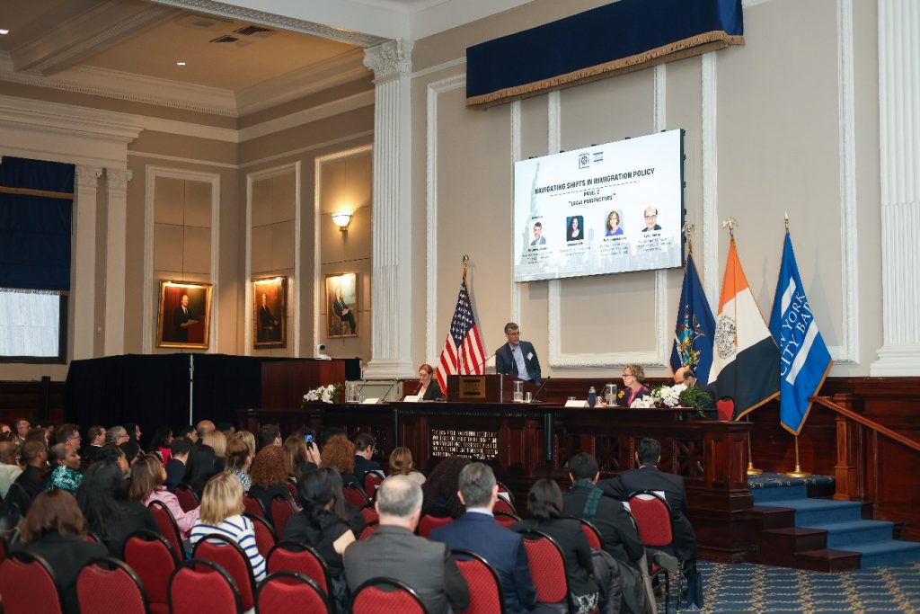 A large seated audience looks up at a raised wooden desk, where a man standing in the center speaks to two women and a man seated next to him, below a screen displaying the title "Navigating Shifts in Immigration Policy: Part 2: Legal Perspectives."
