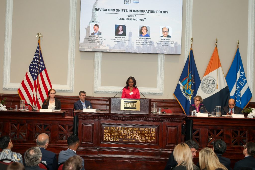 A woman in a pink blouse speaks at the central podium of a raised wooden desk, above a plaque that says "New York City Bar." On either side are four seated people who listen to her speak, as well as several flags.
