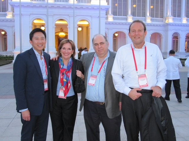 A woman and three men, in professional dress with conference lanyards, pose outside of a white building with a colonnade.