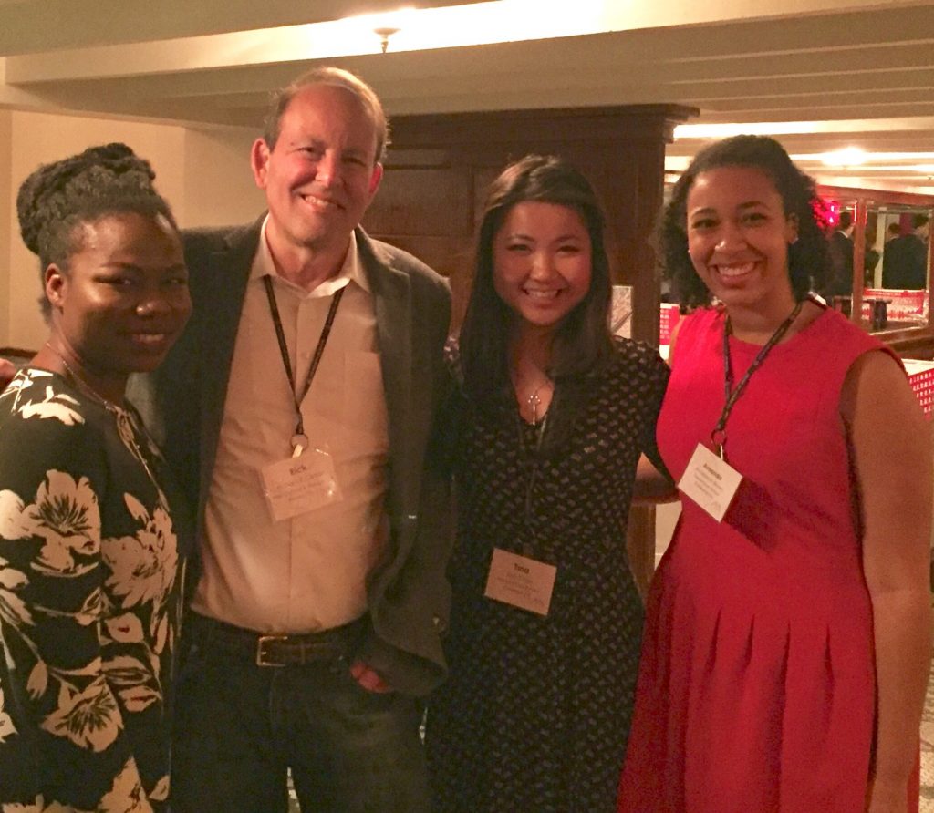 Three women and a man, in professional dress with conference lanyards, pose with arms around each other's backs.