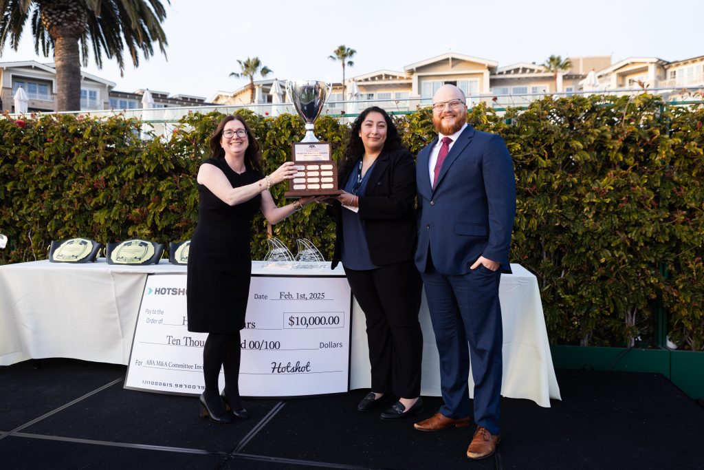 A smiling woman hands a large trophy to a pair of law students, a woman and a man, on a dais outside.